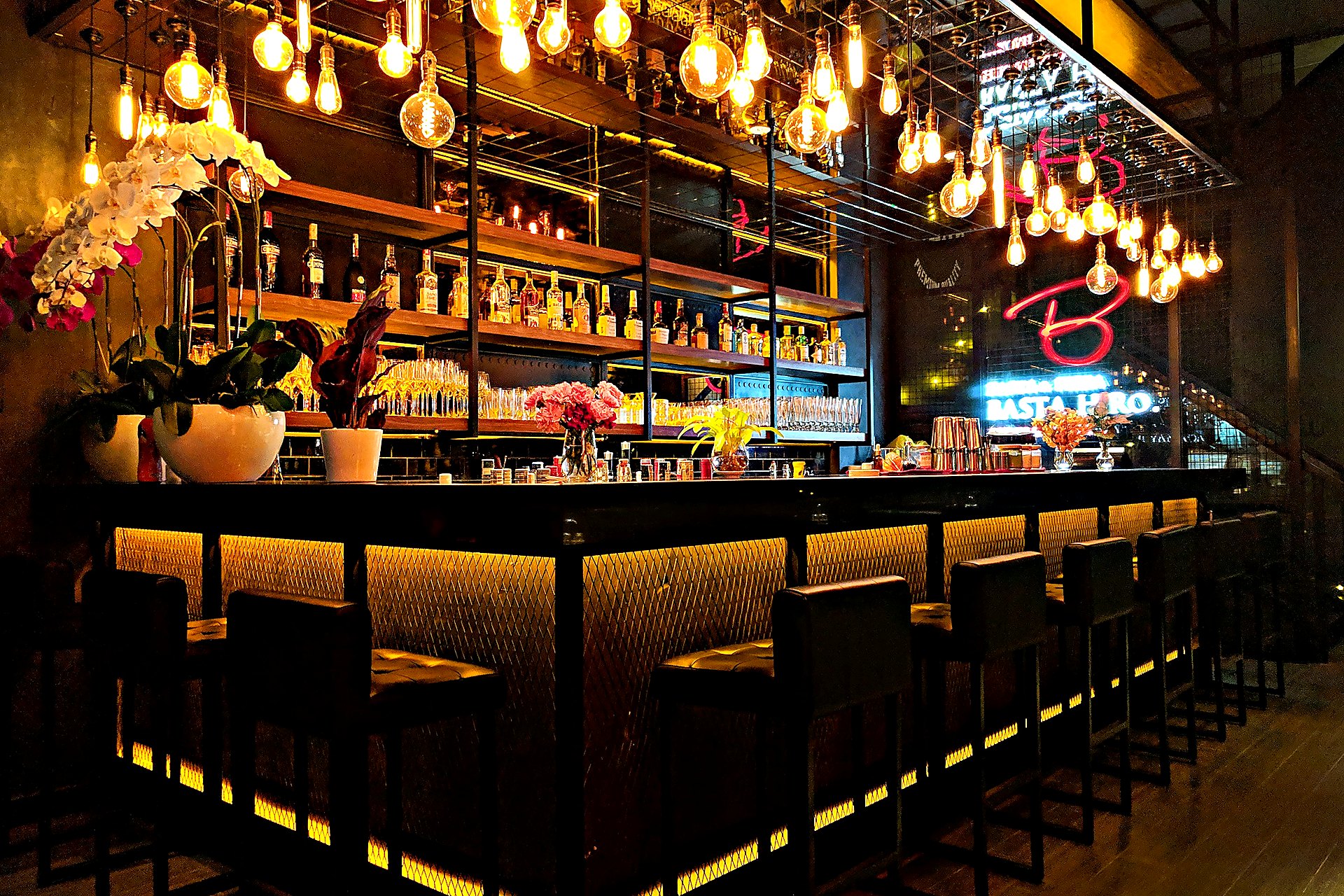 Dim upscale bar with Edison pendant lights, gold under-bar glow, and black leather stools.