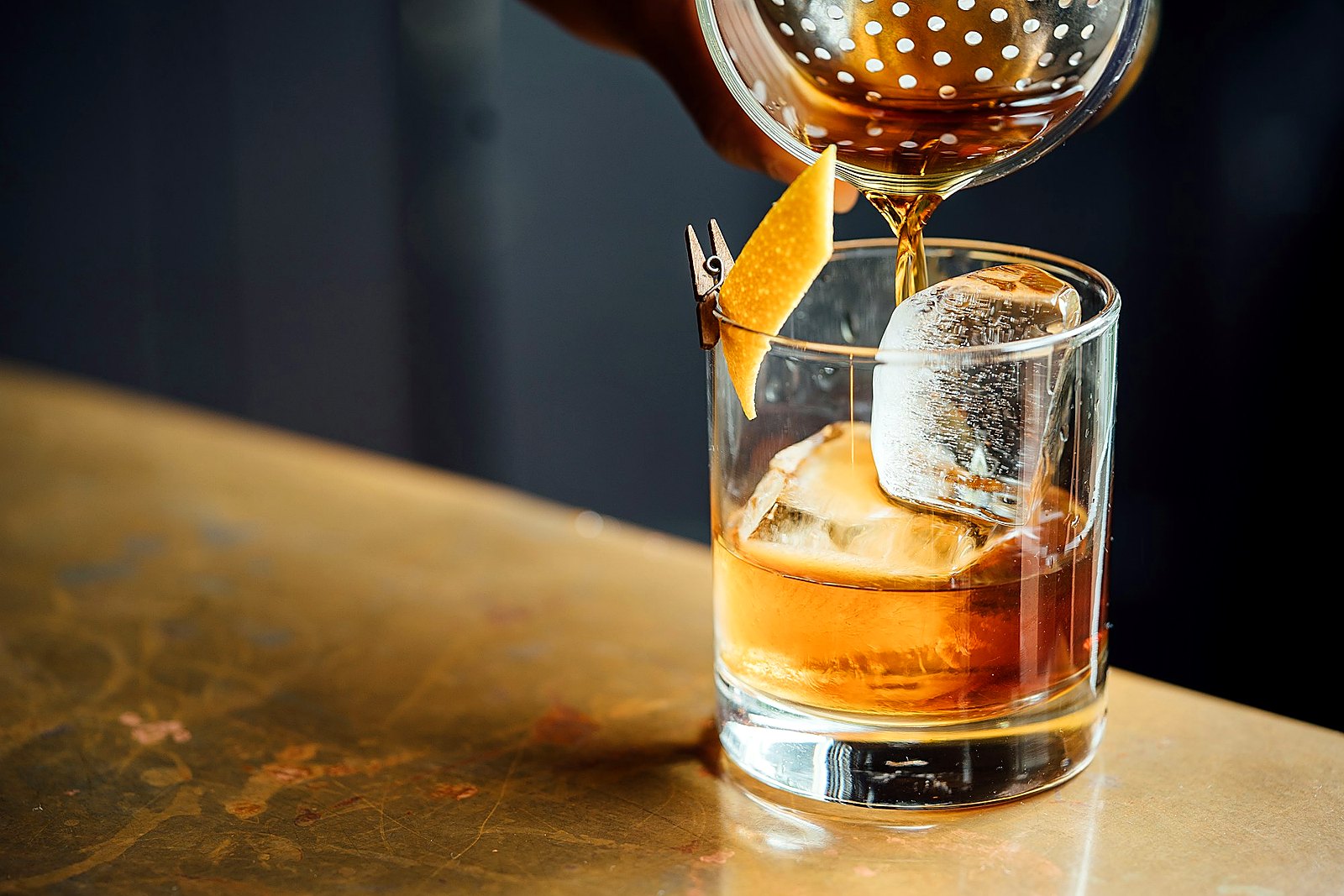 Amber spirit poured through a strainer into a rocks glass with clear ice on a copper bar.