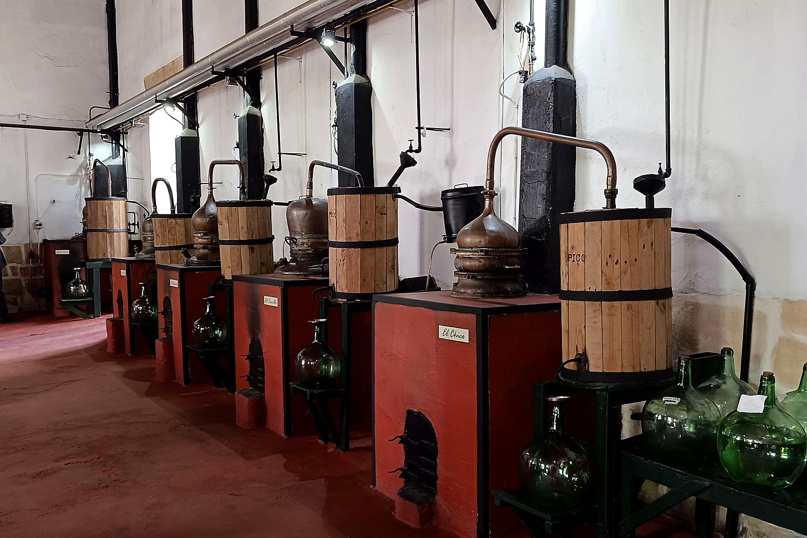 Row of traditional copper alembic stills with wooden condensers in a bright hall.