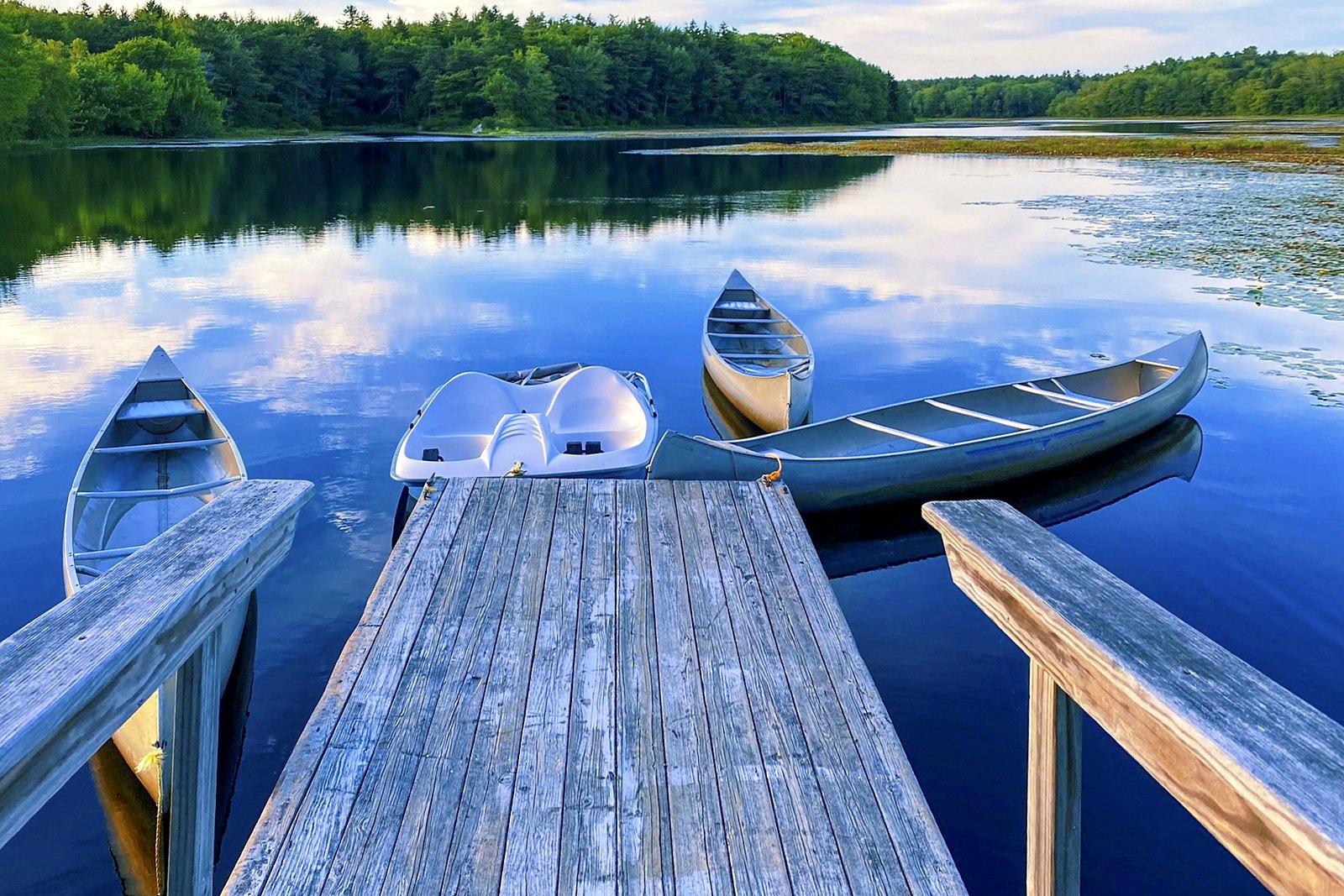 Canoes and a paddleboat moored at the end of a wooden lake dock with mirrored water.