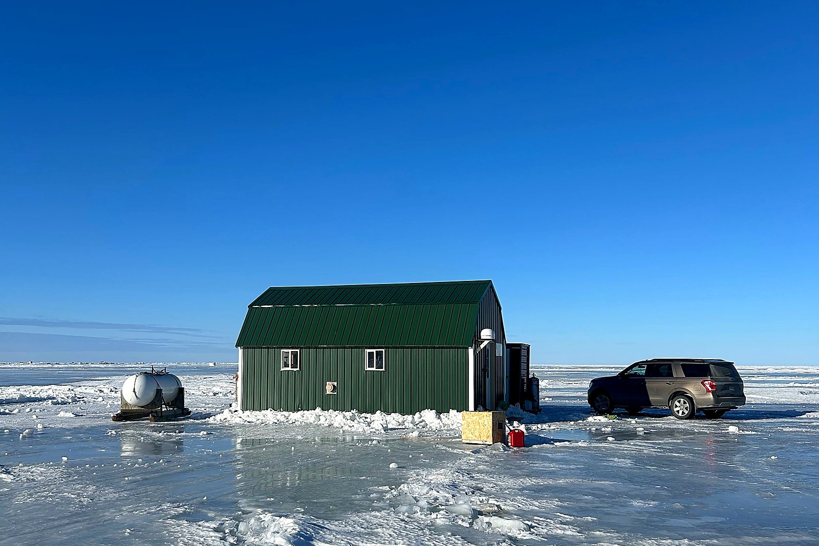 Green ice fishing house on bright hardwater under a clear winter sky.