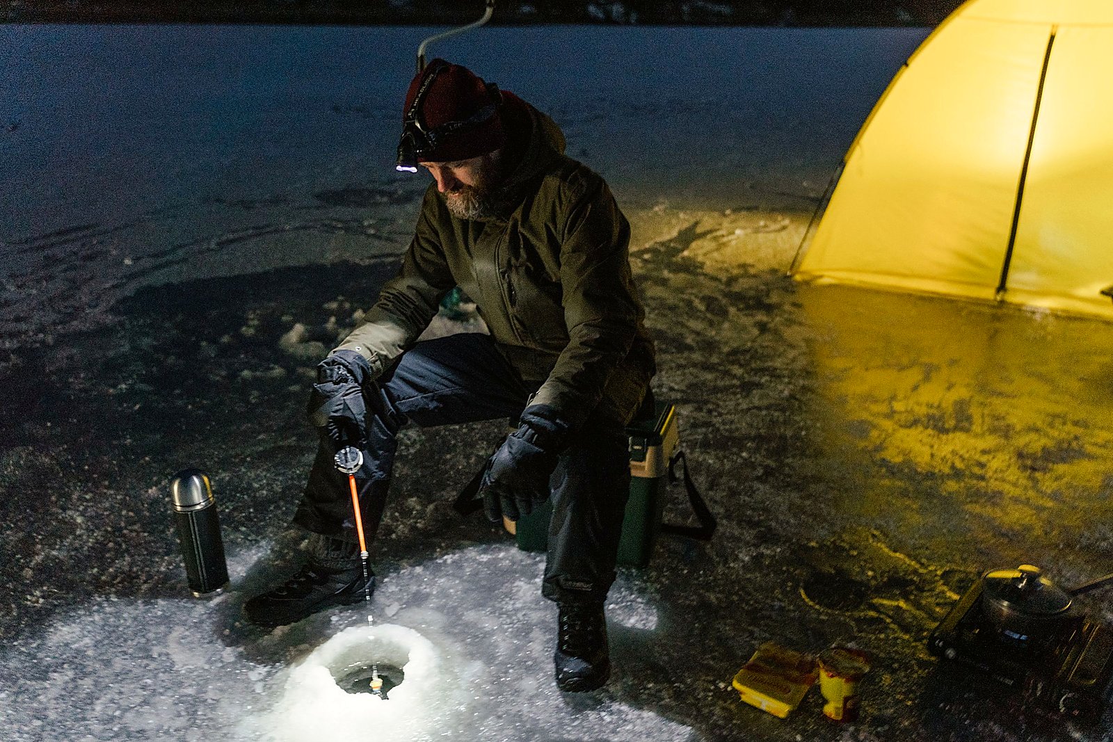 Night ice fishing beside a glowing yellow shanty under a headlamp.