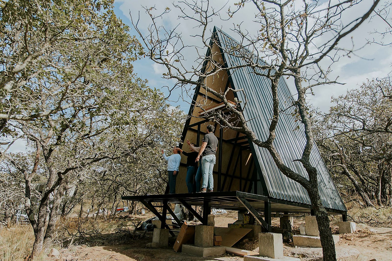 Modern A-frame cabin rising on concrete pier foundations in a wooded clearing.