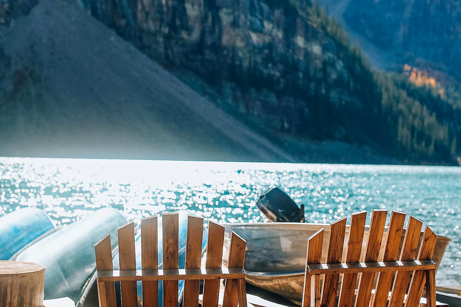 Two wooden Adirondack chairs on a sunny dock facing turquoise lake and mountains.