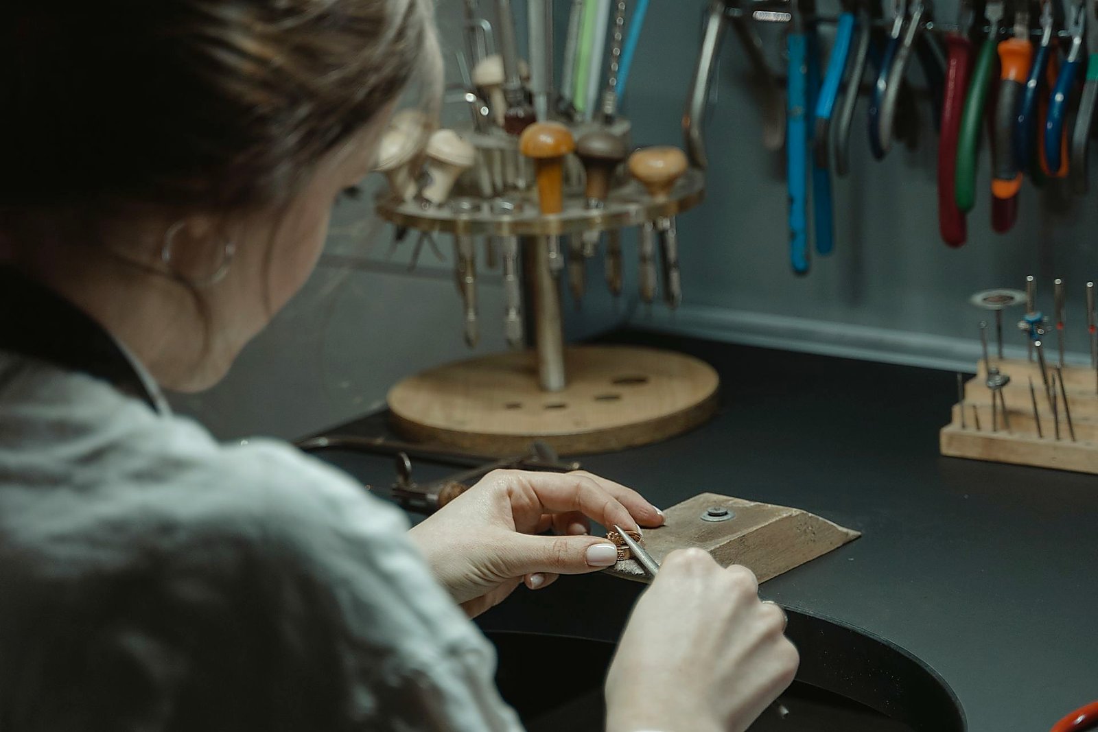 Over-the-shoulder view of a jeweler filing metal at a wooden bench pin.